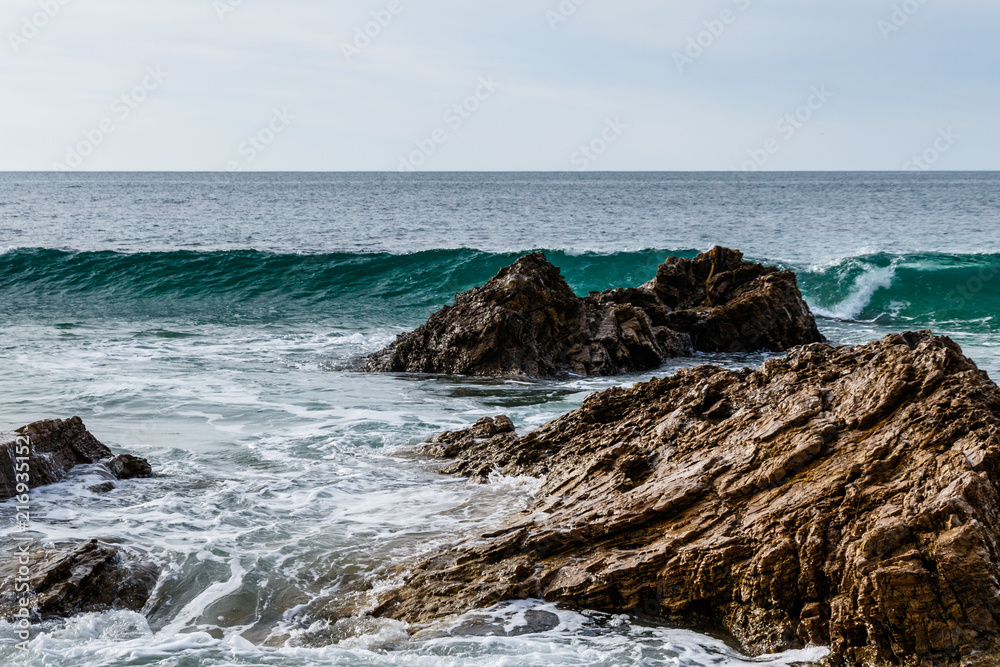 Wave cresting behind rocks near the beach of Crystal Cove State Park in Laguna Beach, California. Foam from an earlier wave is in the foreground; Pacific ocean is in the background.