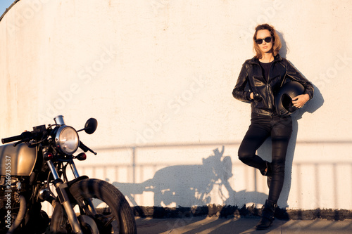 Sexy biker woman in black leather jacket and sunglasses with vintage custom caferacer motorcycle near wall. Urban roof parking, sunset in big city. Traveling and active hipster lifestyle. Girls power.
