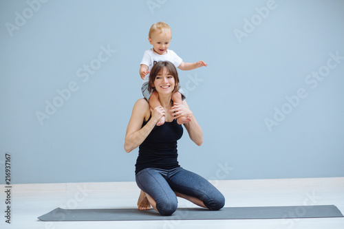 European sportive mother piggybacking her toddler baby son in fitness clothing on gray background. Motherhood, healthy lifestyle concept.