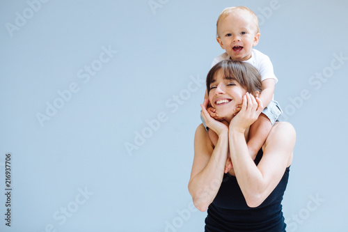 Happy blond toddler boy laughing on to his sportive mother for a piggyback ride over gray background. Close up portrait. Fitness, happy maternity yoga with children concept.
