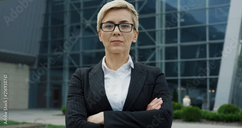 Portrait shot of the beautiful middle aged businesswoman in the glasses with hands crossed in front of her and turning her head to the camera near the big office biuilding. photo