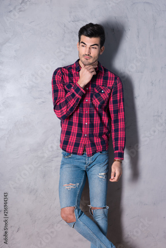 A thinking handsome young man in a red checkered shirt standing in front of a grey wall in a studio.