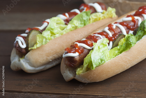 Two homemade Hot Dogs with mayonnaise, ketchup, and green lettuce leaves decorated with baking paper, over wooden table.