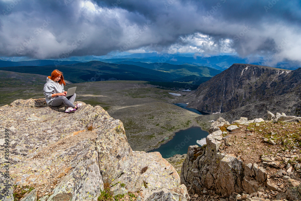 Fototapeta premium Young tourist woman working on a laptop and sitting on the top of the mounting and looking at a beautiful landscape. Freelancer works outside the office. The concept of traveling and working outside