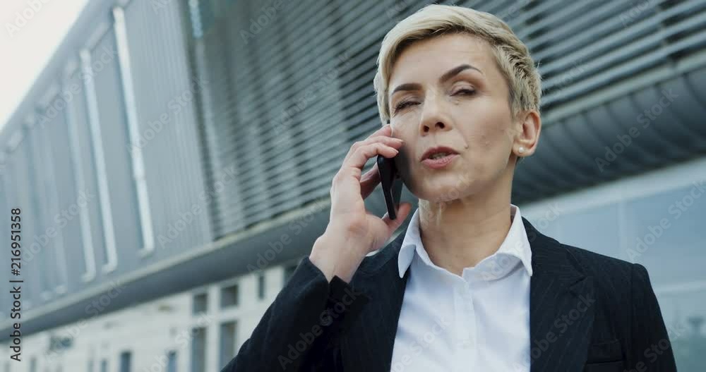 Close up of the Caucasian middle aged serious businesswoman with short blonde hair and in the shirt and jacket having a phone conversation outdoors. Close up of the Caucasian middle aged serious businesswoman with short blonde hair and in the shirt and jacket having a phone conversation outdoors.