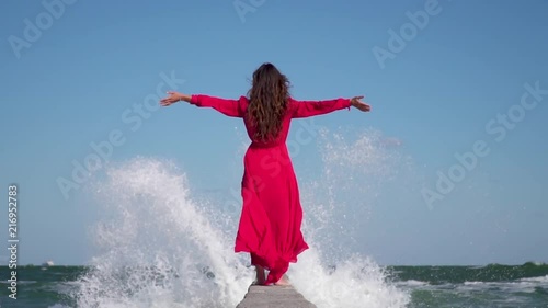 A girl in a red dress is standing on a pier on a background of waves and splashing water