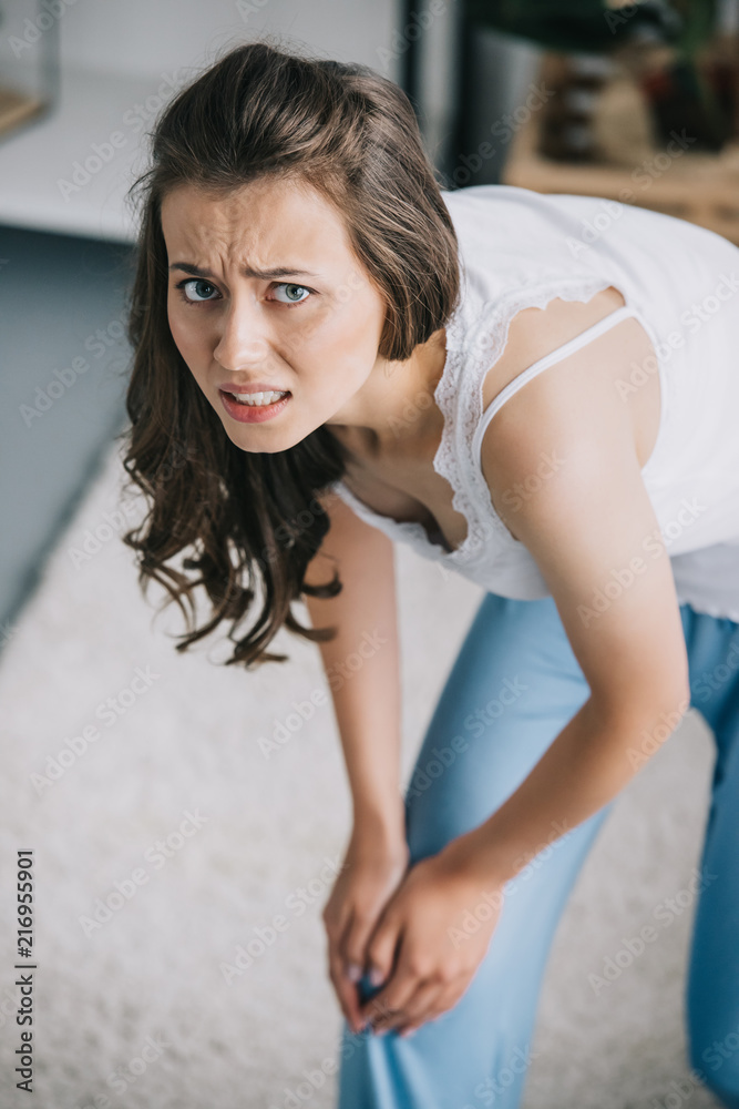 young woman in pajamas suffering from pain in knee and looking at camera