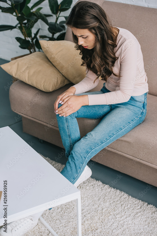 high angle view of woman with pain in knee sitting on sofa at home high angle view of woman with pain in knee sitting on sofa at home