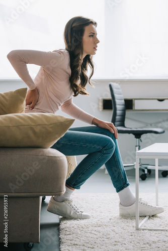 side view of young woman sitting on sofa and suffering from back pain