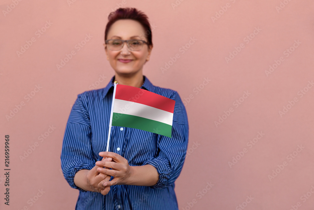 Hungary flag. Woman holding Hungarian flag. Nice portrait of middle aged lady 40 50 years old with a national flag of Hungary over pink wall background.