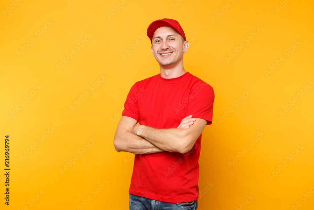 Delivery man in red uniform isolated on yellow orange background. Professional smiling confident male employee in cap, t-shirt courier dealer holding hands crossed folded. Service concept. Copy space.