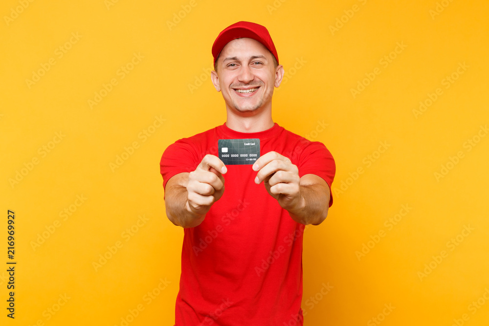 Delivery man in red uniform isolated on yellow orange background. Professional smiling male employee in cap, empty t-shirt courier or dealer hold in hand credit bank card. Service concept. Copy space.