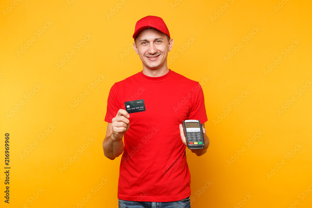 Delivery man in red uniform isolated on yellow background. Male employee in cap, t-shirt courier holding wireless modern bank payment terminal to process and acquire credit card payments, black card. Delivery man in red uniform isolated on yellow background. Male employee in cap, t-shirt courier holding wireless modern bank payment terminal to process and acquire credit card payments, black card.