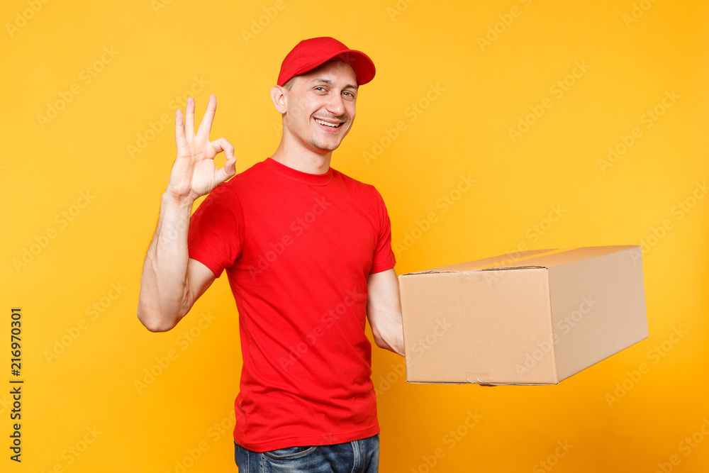 Delivery man in red uniform isolated on yellow orange background. Professional male employee courier or dealer in cap t-shirt holding empty cardboard box. Receiving package service concept. Copy space Delivery man in red uniform isolated on yellow orange background. Professional male employee courier or dealer in cap t-shirt holding empty cardboard box. Receiving package service concept. Copy space