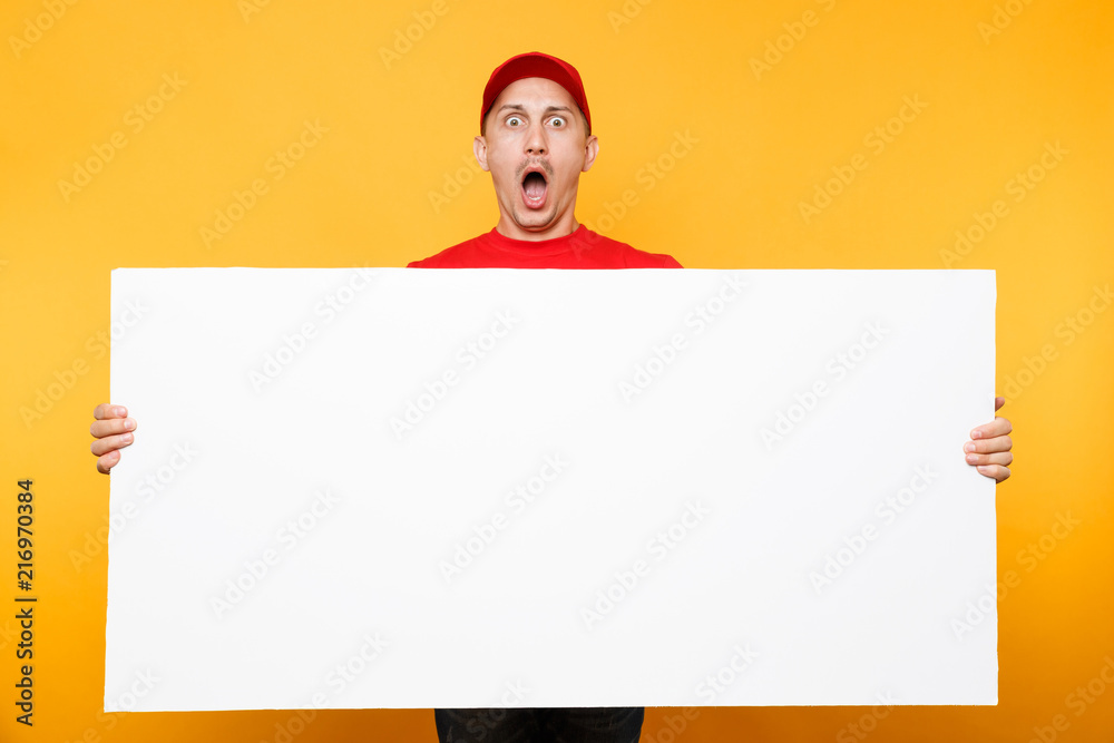 Delivery man in red uniform isolated on yellow orange background. Male employee courier in cap, t-shirt holding big white empty blank billboard with copy space, place for text image. Service concept.