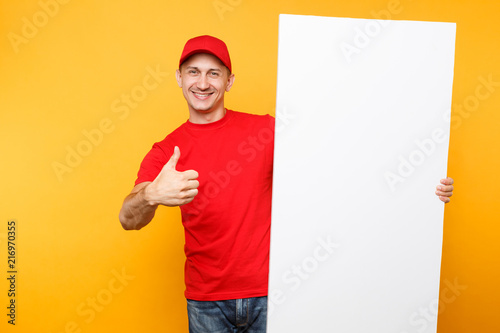 Delivery man in red uniform isolated on yellow orange background. Male employee courier in cap, t-shirt holding big white empty blank billboard with copy space, place for text image. Service concept.