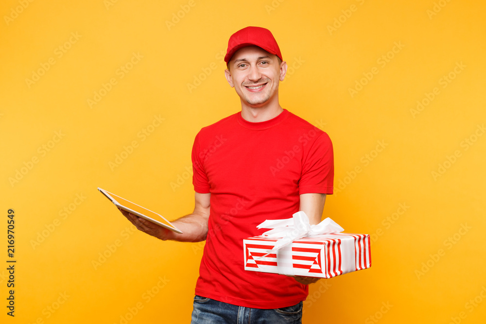 Delivery man in red uniform isolated on yellow orange background. Male employee courier in cap t-shirt holding red present box with ribbon, gift, using tablet pc computer. Service concept. Copy space. Delivery man in red uniform isolated on yellow orange background. Male employee courier in cap t-shirt holding red present box with ribbon, gift, using tablet pc computer. Service concept. Copy space.
