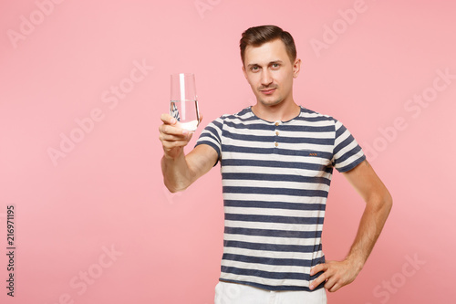 Portrait of smiling young man wearing striped t-shirt holding and drinking clear fresh pure water from glass isolated on trending pastel pink background. People sincere emotions lifestyle concept.