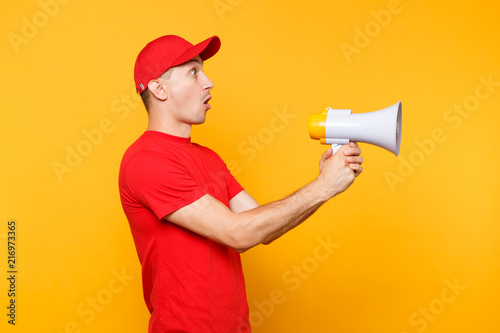 Delivery service man in red uniform isolated on yellow background. Male employee courier in cap, t-shirt screaming in megaphone hot news. Fun guy announces discounts sale. Copy space for advertisement