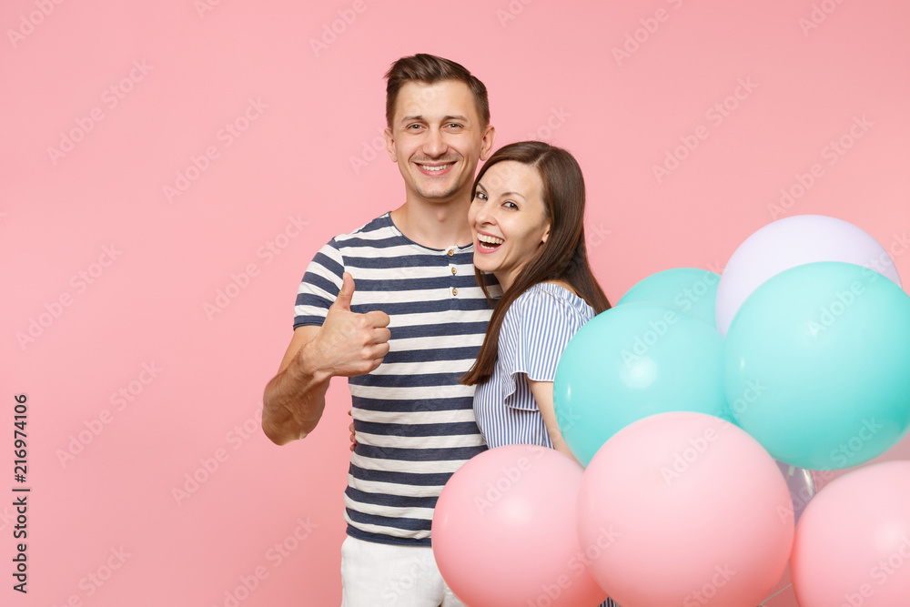 Portrait of young happy smiling couple in love. Woman and man in blue clothes celebrating birthday holiday party on pastel pink background with colorful air balloons. People sincere emotions concept.