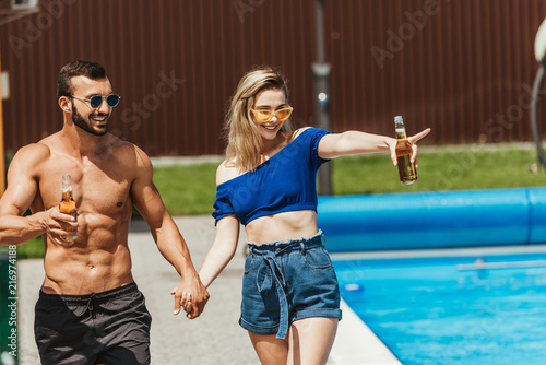 happy couple with bottles of beer holding hands, girl pointing at poolside