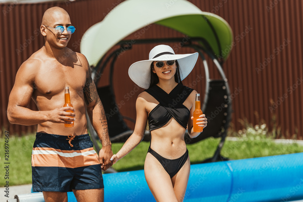 multiethnic couple with bottles of beer and holding hands at poolside