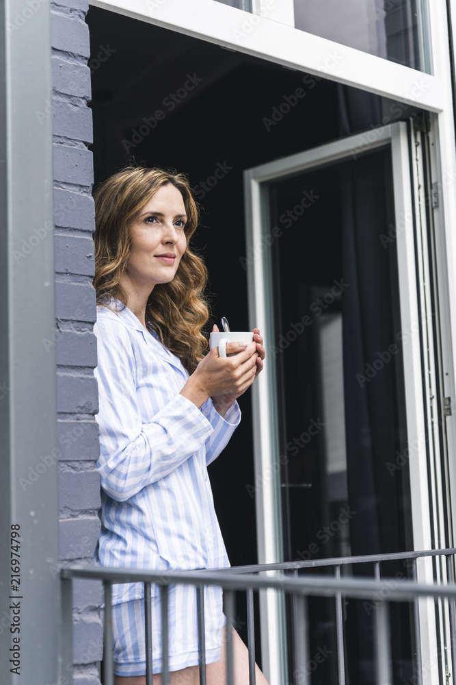 Smiling woman in pajamas at home with cup of coffee looking out of balcony door Smiling woman in pajamas at home with cup of coffee looking out of balcony door