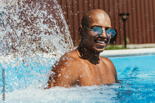 smiling african american man in sunglasses standing in swimming pool with splashes © LIGHTFIELD STUDIOS