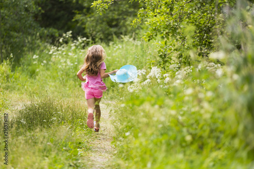 Девочка с длинными светлыми волосами в розовых шортах и футболке с голубой соломенной шляпой в руке бежит в высокой траве