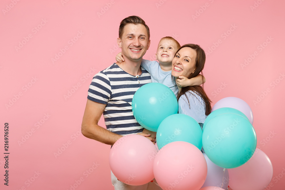 Portrait of young happy family, parents keep in arms, kissing hugging child kid son baby boy, celebrating birthday holiday party on pink background with colorful air balloons. Sincere emotions concept Portrait of young happy family, parents keep in arms, kissing hugging child kid son baby boy, celebrating birthday holiday party on pink background with colorful air balloons. Sincere emotions concept