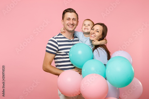 Portrait of young happy family, parents keep in arms, kissing hugging child kid son baby boy, celebrating birthday holiday party on pink background with colorful air balloons. Sincere emotions concept
