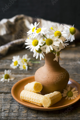 Still life of a bouquet of daisies in a vase and waffle tubes. Wooden rustic style.