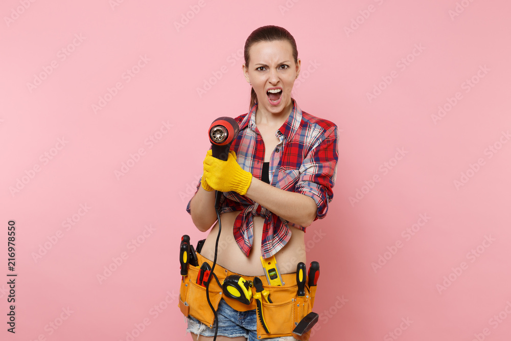 Young beautiful fun handyman woman in plaid shirt, denim shorts, kit tools belt full of variety instruments holding power heat gun isolated on pink background. Female in male work. Renovation concept. Young beautiful fun handyman woman in plaid shirt, denim shorts, kit tools belt full of variety instruments holding power heat gun isolated on pink background. Female in male work. Renovation concept.