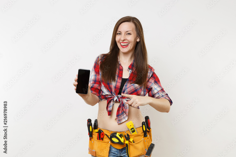 Woman in kit tools belt full of instruments holding showing camera mobile phone with blank black empty screen display touchscreen isolated on white background. Female in male work. Renovation concept. Woman in kit tools belt full of instruments holding showing camera mobile phone with blank black empty screen display touchscreen isolated on white background. Female in male work. Renovation concept.