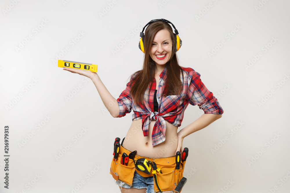 Pretty handyman woman in plaid shirt, denim shorts, noise insulated headphones, kit tools belt full of instruments hold building bubble spirit level isolated on white background. Female in male work. Pretty handyman woman in plaid shirt, denim shorts, noise insulated headphones, kit tools belt full of instruments hold building bubble spirit level isolated on white background. Female in male work.