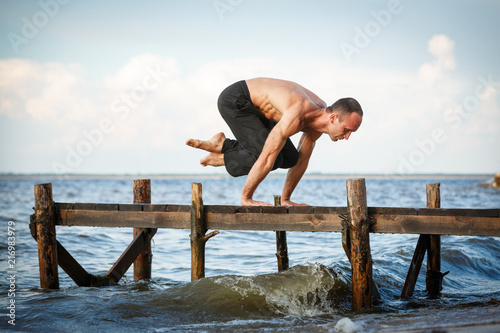 Young yoga trainer practicing lolasana pose on a wooden pier on a sea or river shore
