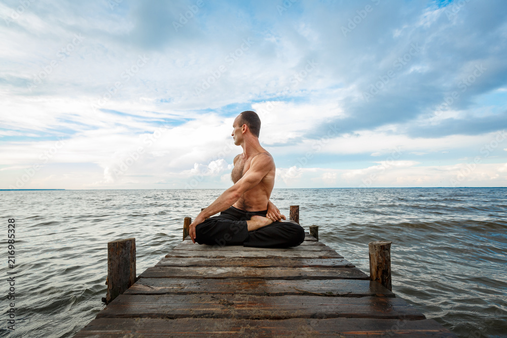Young yoga trainer practicing yoga exercises on a wooden pier on a sea or river shore