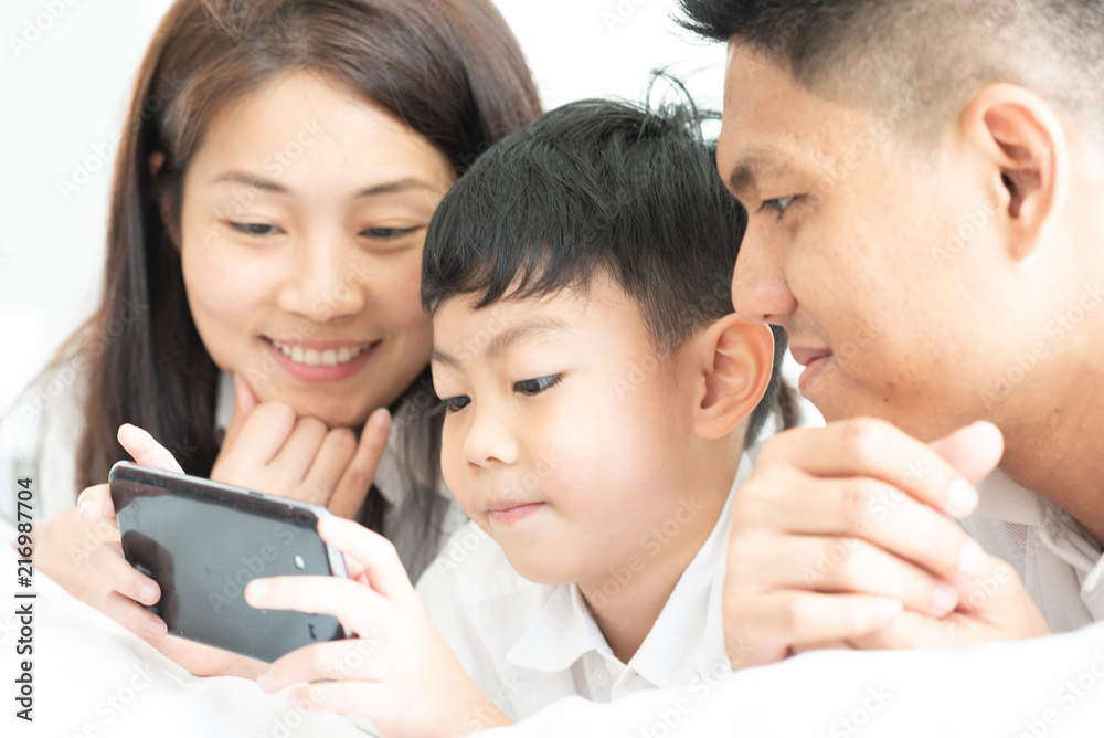 Asian boy playing smartphone with young parents lying in bed.Happy famility Asian boy playing smartphone with young parents lying in bed.Happy famility