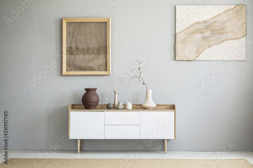 Front view of burlap artworks on a light gray wall above a wooden cupboard with decorations in real photo of a living room interior with a place for your armchair