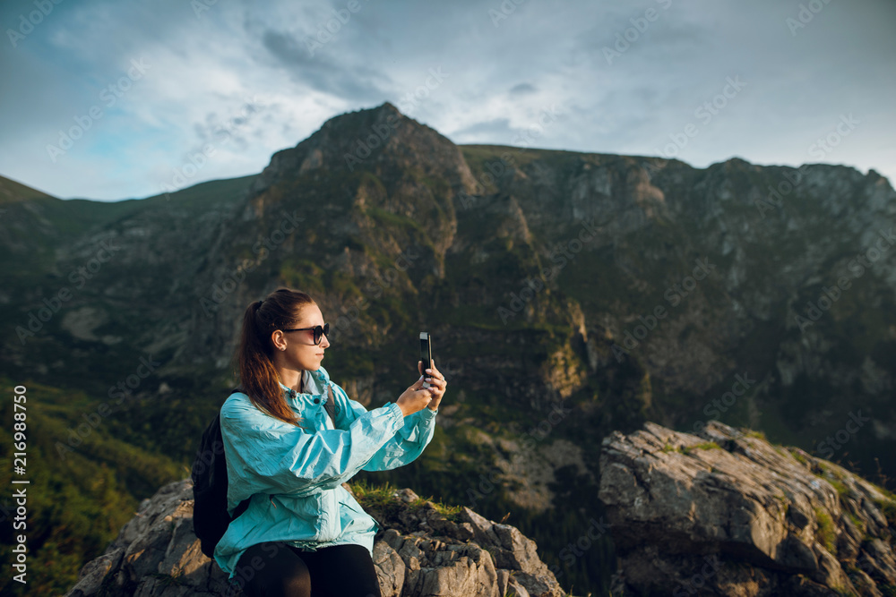 Beautiful young woman in blue jacket, with backpack sitting on the rocks on the Rysy mountain peak in Tatras, Slovakia and Poland ans taking photos on the smartphone.