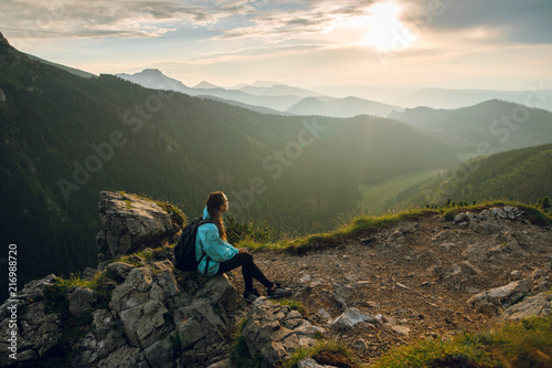 Side view shot of beautiful young woman in blue jacket, with backpack sitting on the rocks on the Rysy mountain peak in Tatras, Slovakia and Poland. Beautiful sunset