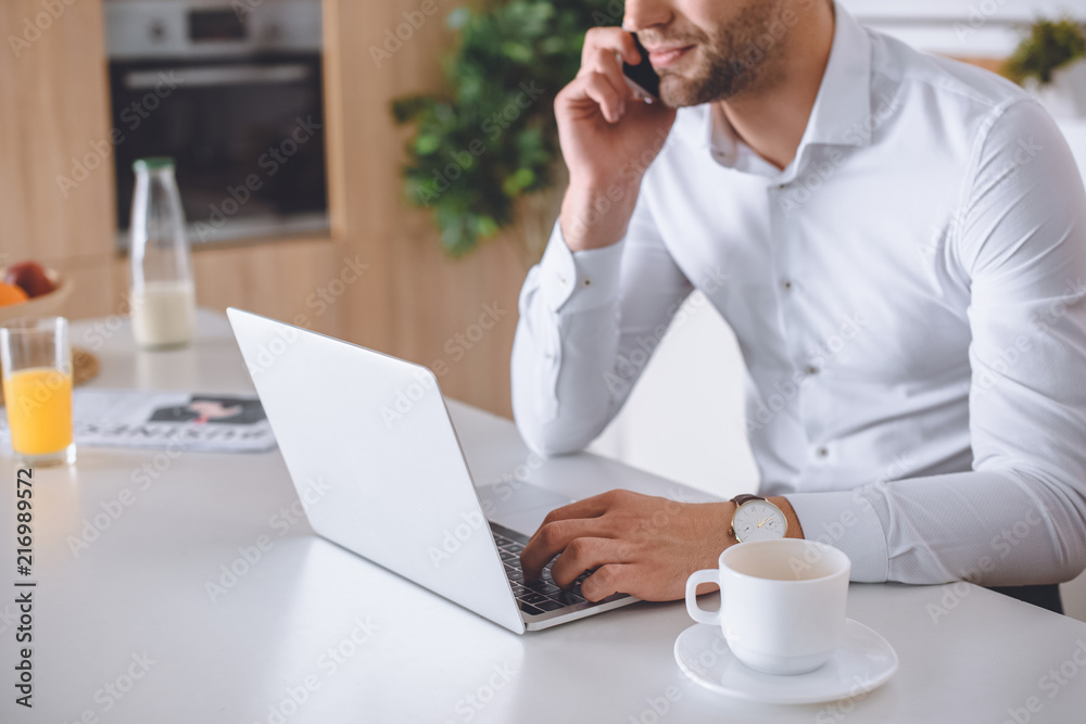 cropped image of businessman talking on smartphone and using laptop at kitchen table with coffee cup cropped image of businessman talking on smartphone and using laptop at kitchen table with coffee cup