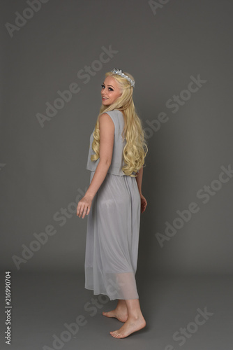 full length portrait of blonde girl wearing dress and crown. standing pose with back to the camera. grey studio background.