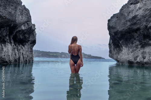 Canvas-taulu Beautiful girl in a black swimsuit in a blue lagoon among the rocks
