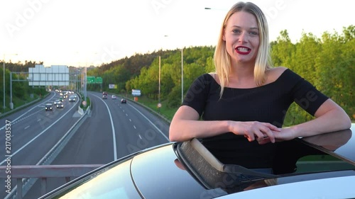 A young beautiful woman stands through a sunroof in a car and talks to the camera with a smile - a highway in the background photo