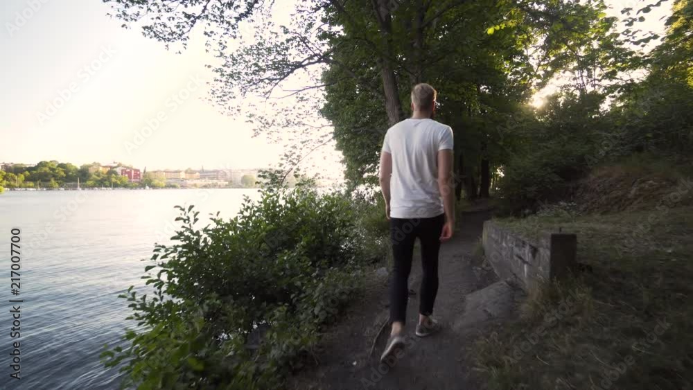 A tall Caucasian male walks by a canal in a forest area in Stockholm Sweden. Beautiful sunny summer evening, green trees and calm ocean. Steady glide cam footage following the model from behind. A tall Caucasian male walks by a canal in a forest area in Stockholm Sweden. Beautiful sunny summer evening, green trees and calm ocean. Steady glide cam footage following the model from behind.