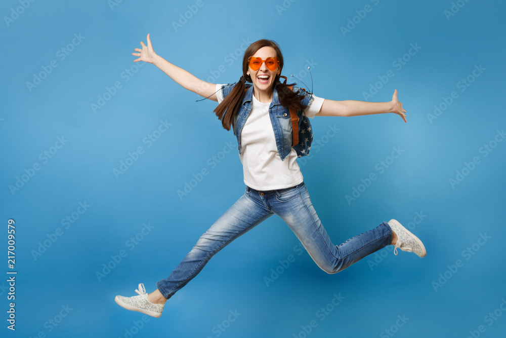 Obraz premium Full length portrait of young overjoyed woman student with backpack in orange heart glasses jump spread hands, legs isolated on blue background. Education in high school. Copy space for advertisement.