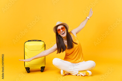 Tourist woman in summer clothes, hat sit at suitcase crossed legs spreading hands as in flight isolated on yellow orange background. Passenger traveling abroad on weekend getaway. Air journey concept.