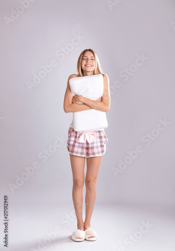 Young woman in pajamas embracing pillow on gray background