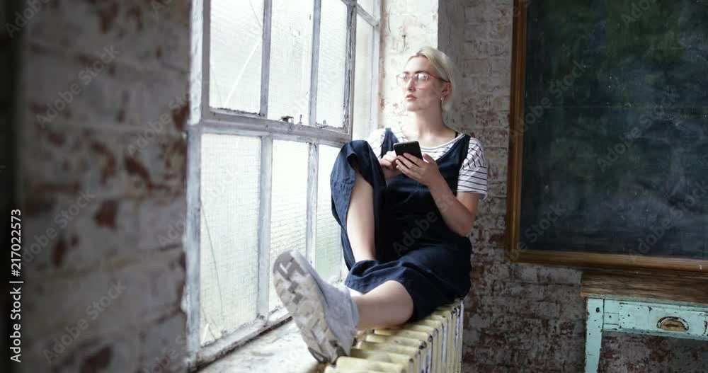 Millennial using smartphone by window in a loft apartment Millennial using smartphone by window in a loft apartment
